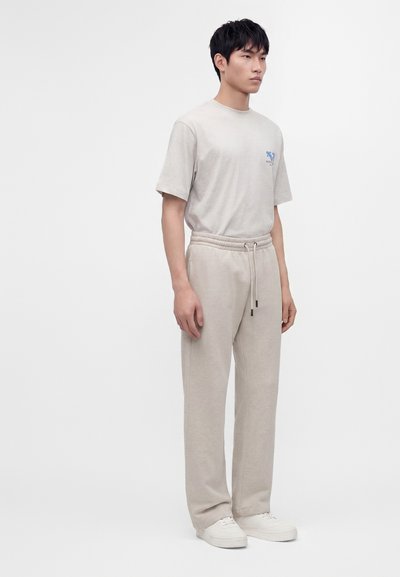 Jeune homme debout portant un t-shirt beige décontracté avec un petit logo bleu, un pantalon à cordon assorti et des baskets blanches sur fond blanc.