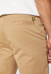 Pantaloni khaki con una trama liscia, dotati di una tasca posteriore e un piccolo logo ricamato giallo sul lato destro della tasca.
