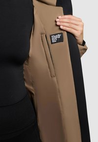 Brązowa i czarna kurtka o gładkiej teksturze, z wewnętrzną kieszenią z metką. Tkanina wydaje się być wodoodporna.