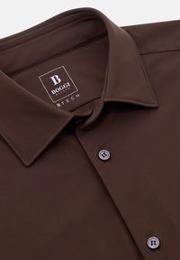 Rudas marškinis su atlenktu apykakle, turintis švelnų tekstūrą ir du priekiniai mygtukai. Vidinėje apykaklėje matomas logotipo etiketė.