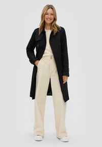 Manteau noir avec de grands boutons, porté sur un haut crème et un pantalon large beige. Tissu lisse avec un design classique et des accents simples.