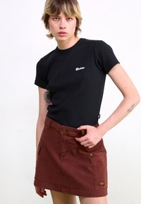 T-shirt di cotone nero con maniche corte, dotato di un logo ricamato bianco. Abbinato a una gonna di denim color ruggine con tasche.