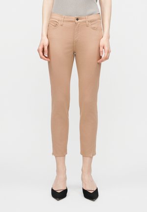 Frau trägt beigefarbene, eng anliegende Knöchelhosen und schwarze spitze High Heels, steht vor einem schlichten hellen Hintergrund.