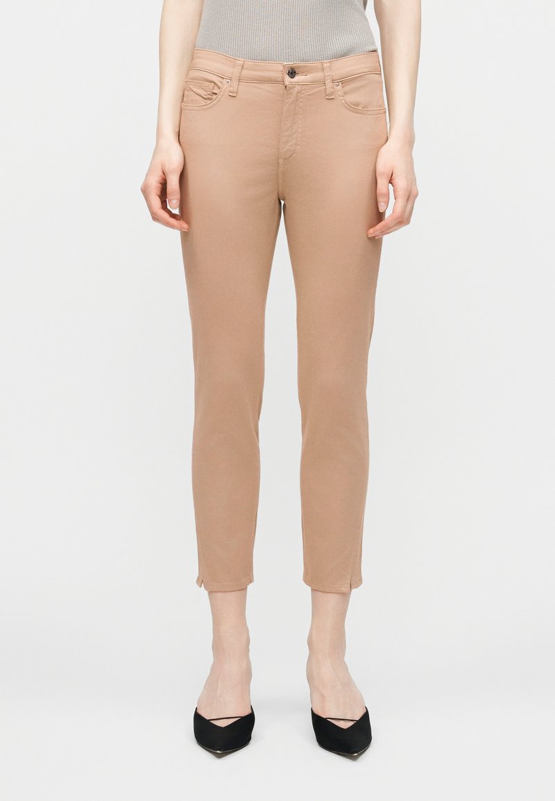 Γυναίκα που φορά μπεζ στενά παντελόνια μέχρι τον αστράγαλο και μαύρες γόβες με μυτερή μύτη, στέκεται μπροστά σε απλό φωτεινό φόντο.