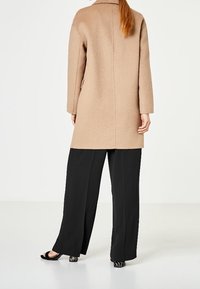 Abrigo de lana beige con mangas largas, silueta recta y detalle de costura en la parte trasera. Combinado con pantalones negros holgados y calzado de punta abierta.