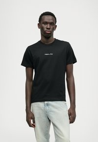 Svart bomulls t-skjorte med rund hals og korte ermer, med hvit logotekst "REPLAY" sentrert på brystet. Brukt med lyseblå jeans.