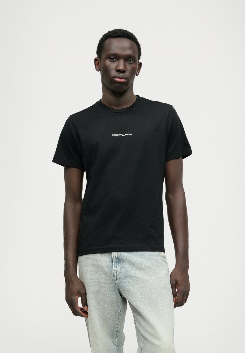 Svart bomulls t-skjorte med rund hals og korte ermer, med hvit logotekst "REPLAY" sentrert på brystet. Brukt med lyseblå jeans.