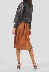 Zwarte leren jas met een aansluitend ontwerp, gecombineerd met een vloeiende, enkel-lange bruine jurk. De jurk heeft een subtiele glans en geplooide details. Zwarte hoge hakken maken de outfit compleet.