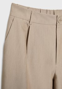 Pantalons beige ajustés en tissu lisse, avec un devant plissé et une fermeture à bouton à la taille. Poche fonctionnelle sur le côté.