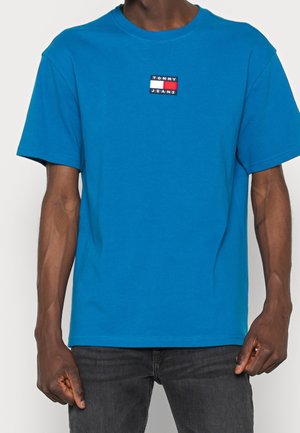 Mies, jolla on sininen lyhythihainen Tommy Jeans -t-paita, jossa rintamuksessa logoplässi, ja tummanharmaat housut, seisoo rentoutunein käsin.