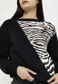 Felpa nera in cotone con un motivo diagonale a strisce bianche di zebra su un lato. Polsini e orlo a costine, con una vestibilità rilassata.
