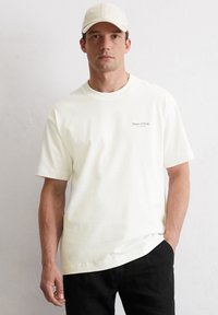 White cotton t-shirt with a crew neckline and short sleeves, featuring a small black logo text "Marc O'Polo" on the front. Worn with black pants.