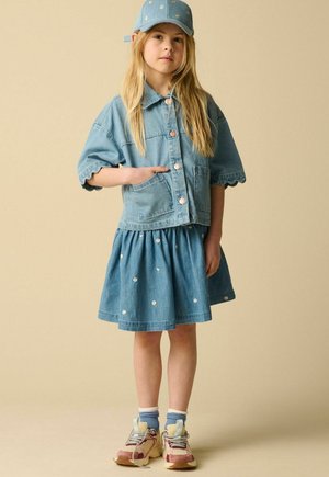 Young girl wearing denim jacket, floral skirt, matching cap, and sneakers, standing with one hand in pocket against beige background.