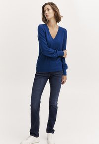 Modra V-izrezana pulover iz mehkega pletenega materiala, z dolgimi rokavi in sproščenim krojem, v kombinaciji s temnimi naravnost krojenimi kavbojkami in belimi supergami.