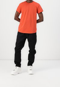 Orange bomullströja med korta ärmar, rund hals, kombinerad med svarta elastiska byxor och vita sneakers med texturerade sulor.