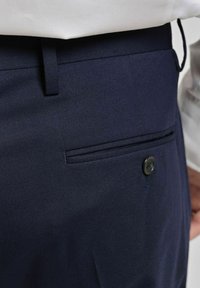 Marinblå byxor i texturerat tyg, med en bakficka, stängning med svart knapp och bälteshällor i midjan.