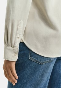 Ljusbeige kord skjorta med knappmanschetter, med rundad nederdel. I kombination med mörkblå denimjeans med klassisk fickdesign.