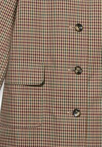 Giacca doppiopetto beige, con un piccolo motivo a quadri in bordeaux e verde. Include una tasca sul petto e bottoni rotondi.