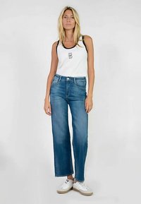 Camiseta blanca sin mangas con ribete negro; pantalones vaqueros azules de pierna ancha; zapatillas blancas con suelas beige. Conjunto informal, sin patrones o adornos visibles.