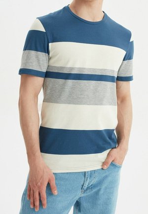 Man draagt een T-shirt met korte mouwen en een ronde hals, voorzien van brede horizontale strepen in blauw, wit en grijs, gecombineerd met lichtblauwe spijkerbroek.