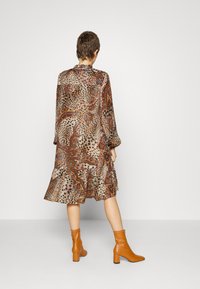 Un mannequin aux cheveux courts porte une longue robe à motifs dans des tons terreux et des bottines orange, se tenant de dos contre un fond uni.