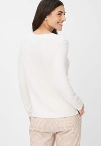 Weißes, langärmliges, geripptes Oberteil mit rundem Halsausschnitt; glatte Textur und dezente Nähtdetails, kombiniert mit hellkarierten Hosen.