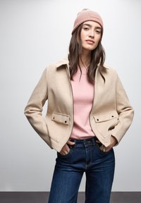Giacca beige con zip e colletto, dotata di due tasche frontali, abbinata a un maglione rosa chiaro e jeans blu scuro, completata da un berretto rosa chiaro.