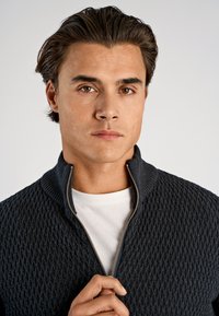 Dunkelgrauer Strickpullover mit Reißverschluss, strukturiertem Rautenmuster, hohem Kragen und silbernem Reißverschluss, getragen über einem weißen Rundhalsausschnitt-Shirt.