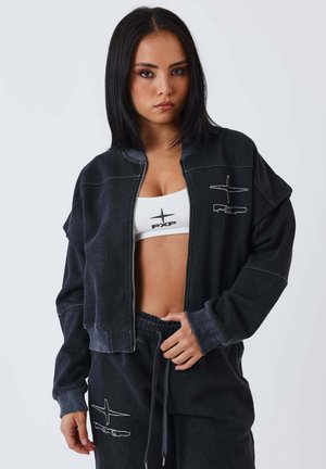 Giovane donna con capelli neri lisci indossa giacca nera con cerniera, pantaloni abbinati e reggiseno sportivo bianco con logo "PXP", posa contro uno sfondo bianco.