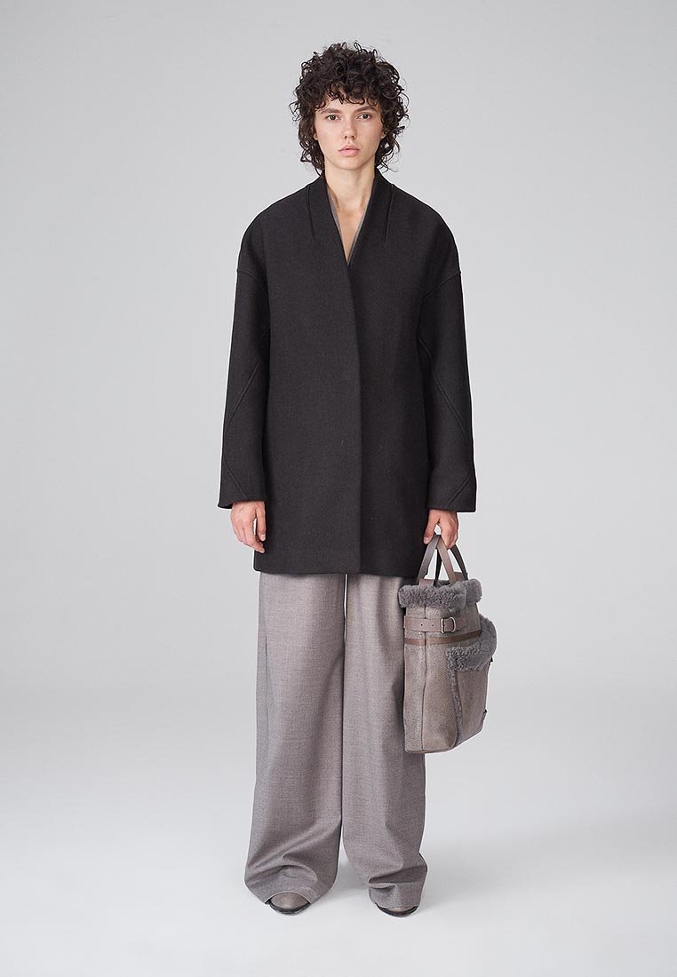 Cappotto nero oversize con scollo a V e maniche ampie, abbinato a pantaloni grigi chiari a gamba larga. Borsa grigia con dettagli in pelliccia.