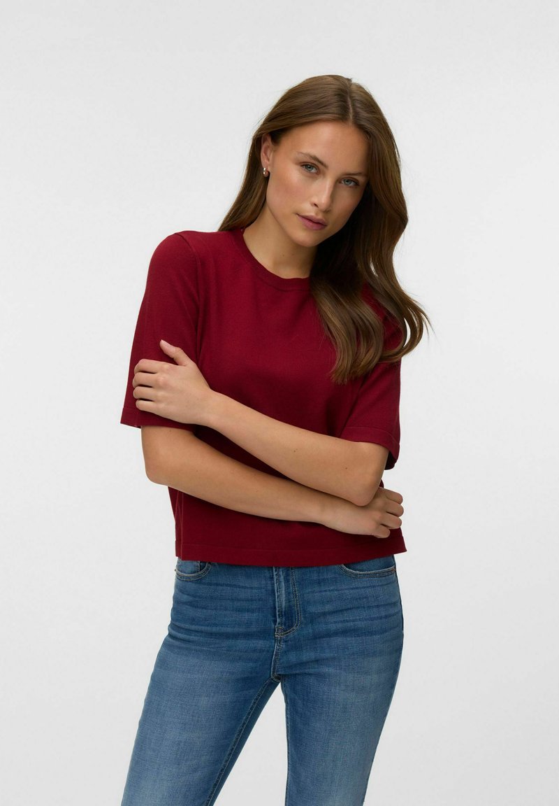 Jeune femme aux longs cheveux bruns portant un haut rouge à manches courtes et un jean bleu, debout les bras croisés devant un fond uni.
