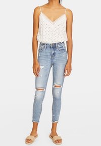 Top blanco sin mangas con lunares negros, jeans ajustados de mezclilla desgastados de color azul claro y sandalias slip-on texturizadas en color beige.