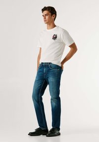 Joven con camiseta blanca con diseño gráfico, jeans azules, zapatos negros, de pie con una mano en el bolsillo, mirando hacia un lado contra un fondo liso.