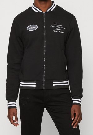 Personne portant une veste noire zippée avec un col, des poignets et un ourlet à rayures blanches, ainsi qu'un texte brodé blanc sur la poitrine.