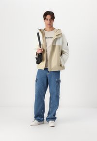 Jachetă crem și albă cu fermoar, pantaloni cargo din denim, geantă crossbody neagră. Modelul poartă un tricou deschis cu logo-ul "Calvin Klein" și adidași albi.