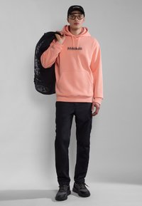 Sudadera rosa claro con bolsillo frontal y logotipo, combinada con pantalones cargo negros y zapatos de senderismo negros. Hombre sosteniendo una mochila negra.