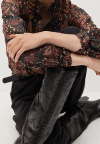 Stivali neri al ginocchio con intricati motivi floreali in rilievo e cuciture texture, abbinati a una blusa a maniche lunghe a fantasia e pantaloni neri.