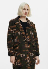 Uldblandet frakke med camouflage mønster i grøn, brun og beige, med en kasketkrave, to frontlommer og en afslappet pasform.