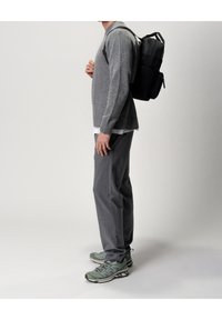 Osoba ubrana w szary sweter, szare spodnie, zielone sneakersy, niosąca czarny plecak, stojąca bokiem na jednolitym tle.