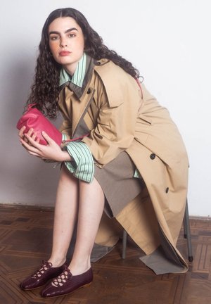 Femme aux longs cheveux bouclés assise sur une chaise, portant un trench-coat beige, une chemise à rayures vertes, tenant une pochette rose et des chaussures à lacets bordeaux.