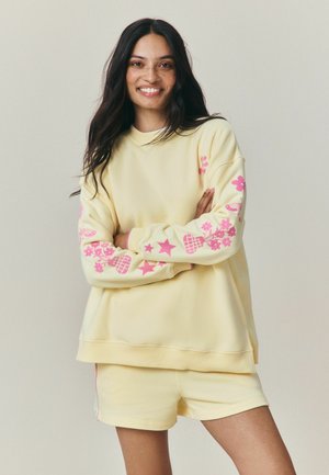 Young woman with long dark hair and freckles wearing a yellow sweatshirt with pink floral patterns and matching yellow shorts smiling with arms crossed.