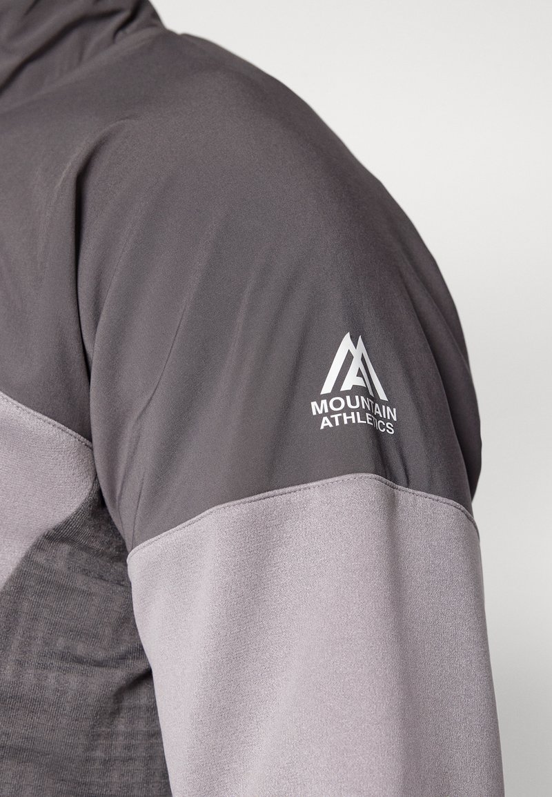 Giacca sportiva grigia caratterizzata da un design strutturato con una parte superiore liscia e il logo "Mountain Athletics" in bianco sulla manica.