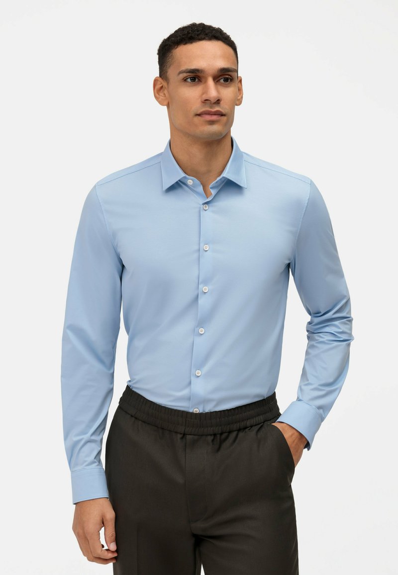 Camicia azzurro chiaro a maniche lunghe con colletto a bottoni e bottoni bianchi, abbinata a pantaloni neri con vita elastica. Tessuto di tipo liscio.