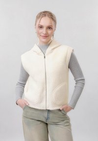 Jeune femme aux cheveux blonds portant un gilet blanc en tissu duveteux zippé par-dessus un pull côtelé gris et un pantalon vert clair, les mains dans les poches, souriante.
