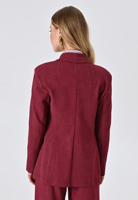 Bordeauxroter Blazer mit glatter Textur, einreihigem Knopfverschluss, Reverskragen und langen Ärmeln, der eine taillierte Passform und einen Rückenbelüftung bietet.