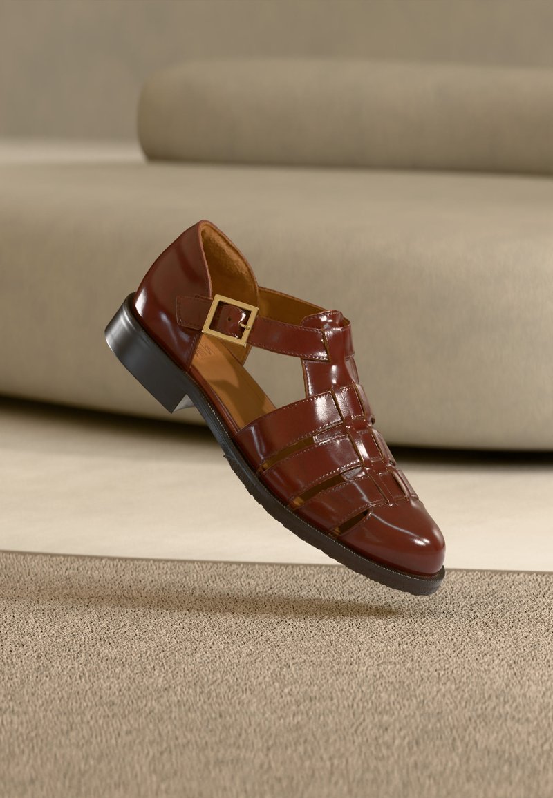 Καφέ δερμάτινο σανδάλι κλειστού τύπου με λουράκι με αγκράφα, μαύρη σόλα και γυαλιστερό φινίρισμα, τοποθετημένο πάνω σε μπεζ χαλί και φόντο καναπέ.