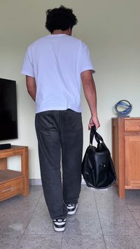 Saco de couro preto com uma forma arredondada e alças duplas. A pessoa veste uma t-shirt branca e calças pretas, estando de pé sobre um chão texturizado.