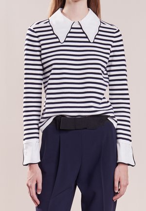 Femme portant une chemise rayée noir et blanc à manches longues avec un grand col blanc et des poignets, associée à un pantalon taille haute bleu foncé.