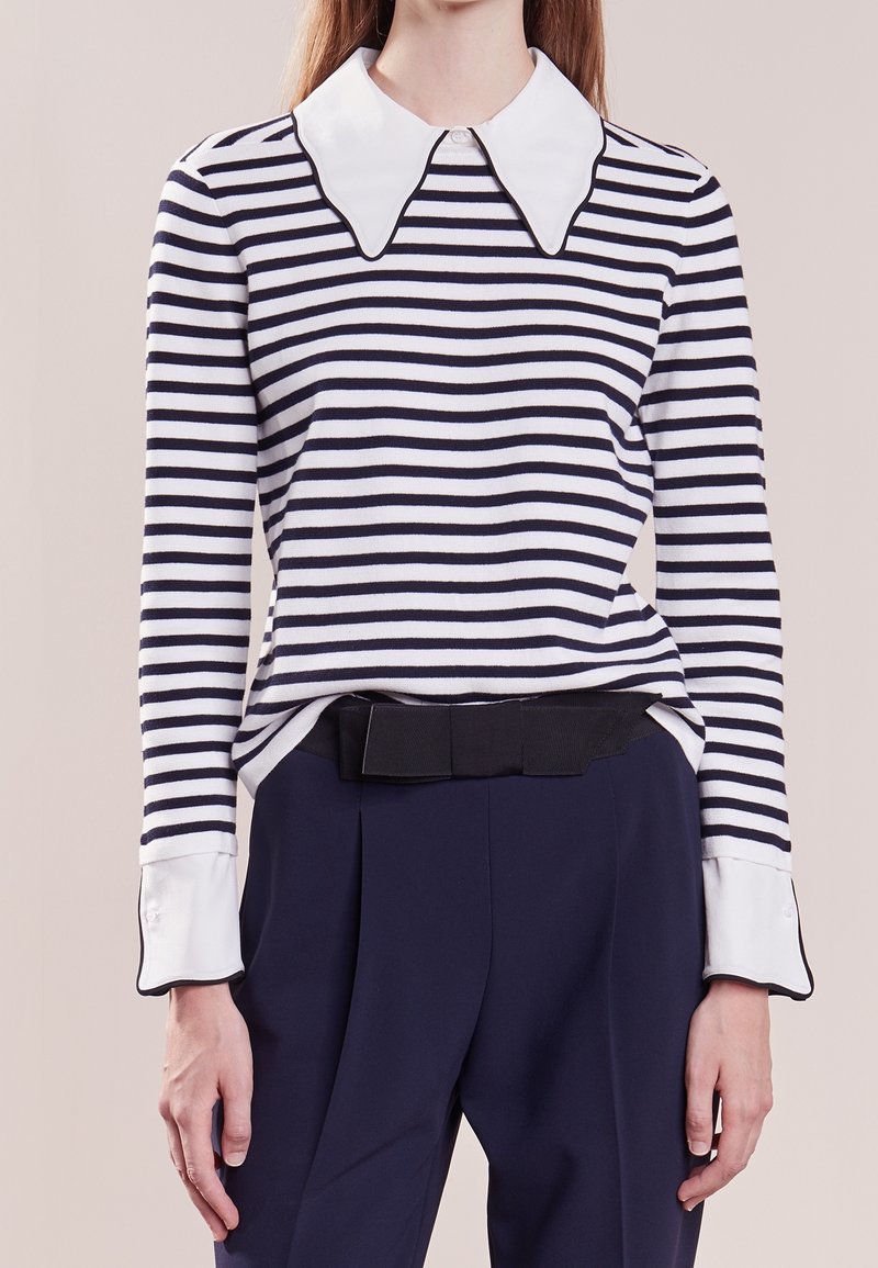 Femme portant une chemise rayée noir et blanc à manches longues avec un grand col blanc et des poignets, associée à un pantalon taille haute bleu foncé.