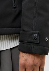 Czarny materiałowy mankiet kurtki z okrągłym czarnym napem. Widoczne szwy wzdłuż krawędzi oraz jasnoszara koszula w paski pod spodem.